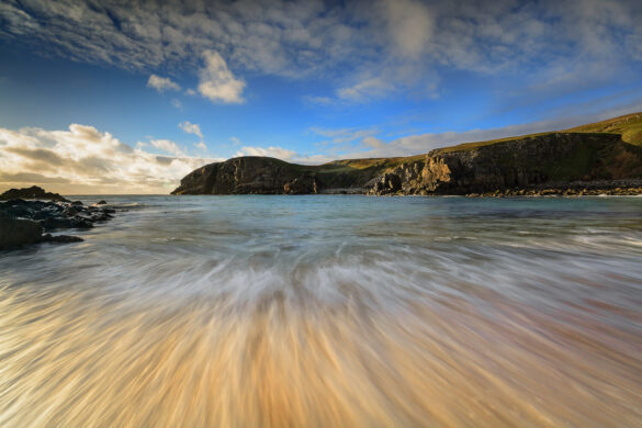 A beach scene where the water is rushing onto the sand, showing a sense of motion, and sky with fluffy white clouds.