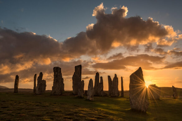 The Callanish Standing Stones at sunset there is a darkening blue sky with orange-rimmed clouds, and the sun is descending behind one of the stones.