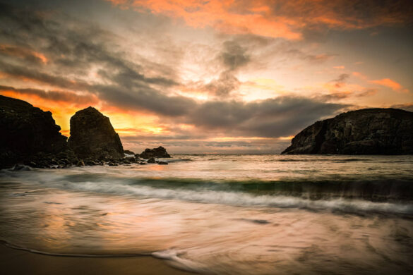 Dalbeg Beach at sunset, the sky is streaked with fiery reds, oranges, and greys, and waves gently curling over rocks in silhouette.