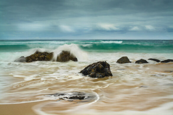 Dalmore Beach with a blue sky and gentle white clouds, aqua marine waves gently curling, and splashing over rocks frozen in time.