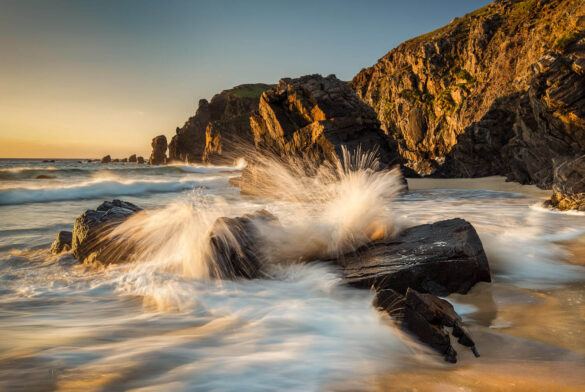 Wave crashing at Dalmore Beach at sunset with golden sunlight on cliff face, water and sand.