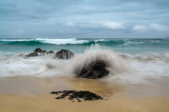 Dalmore Beach with a blue sky and gentle white clouds, aqua marine waves gently curling, and water frozen in time crashing against rocks.