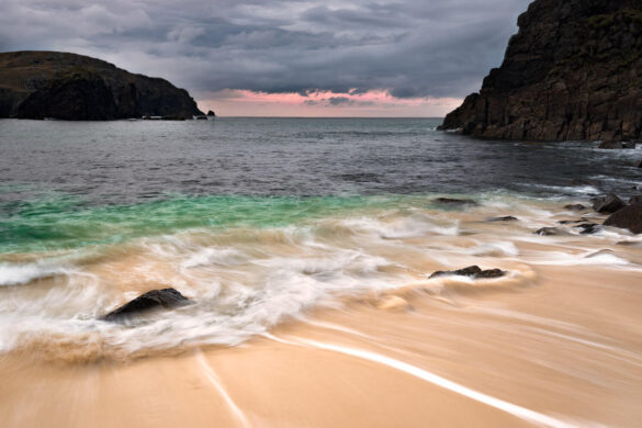 A beach with a wave flowing back and forth, rocks in the foreground, and a mix of aquamarine and inky blue sea.