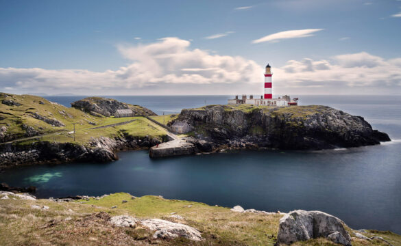 Eilean Glas lighthouse on a rocky promontory, bay stretching into the sea, and a blue sky with streaked white clouds.