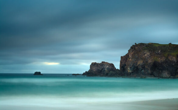 Mangersta Beach with smooth, glassy sea, aquamarine color, and blue sky with white and grey cliffs and rocks.