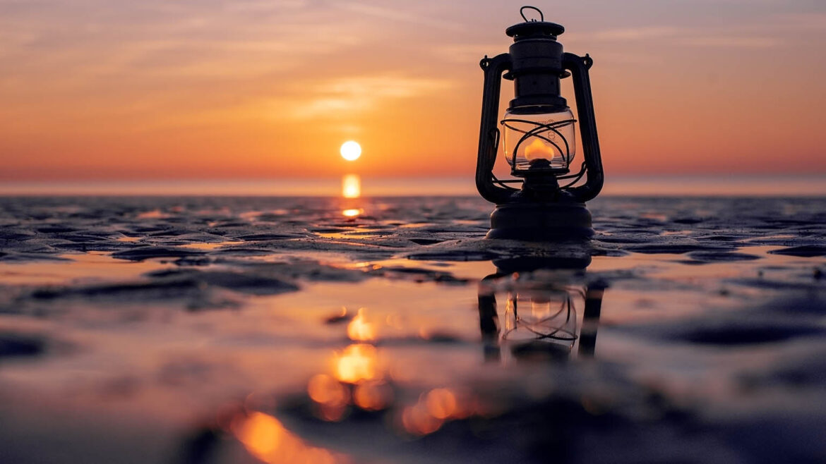 Hurricane lamp with flame on the beach at sunset. Setting sun reflected on the wet sand in the foreground