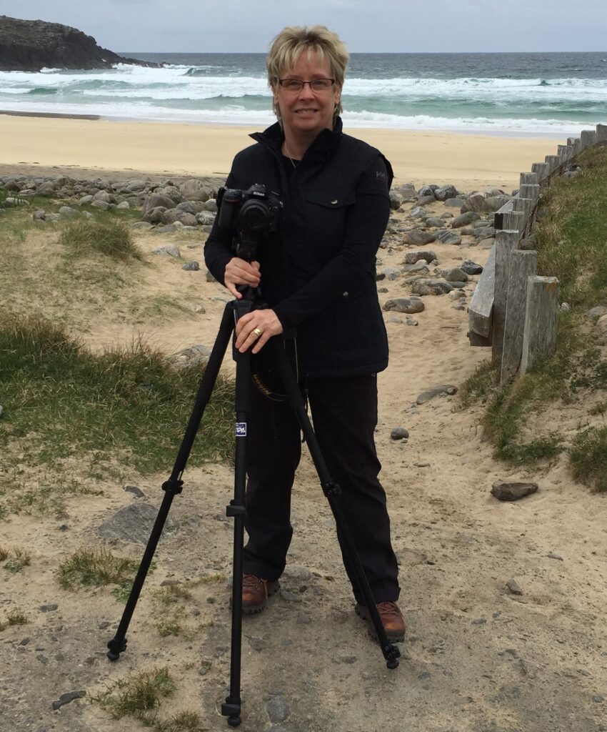 Elaine Cook, known as the Lens Lady, standing with her camera on a tripod at Dalmore Beach, ready for a seascape photo shoot.