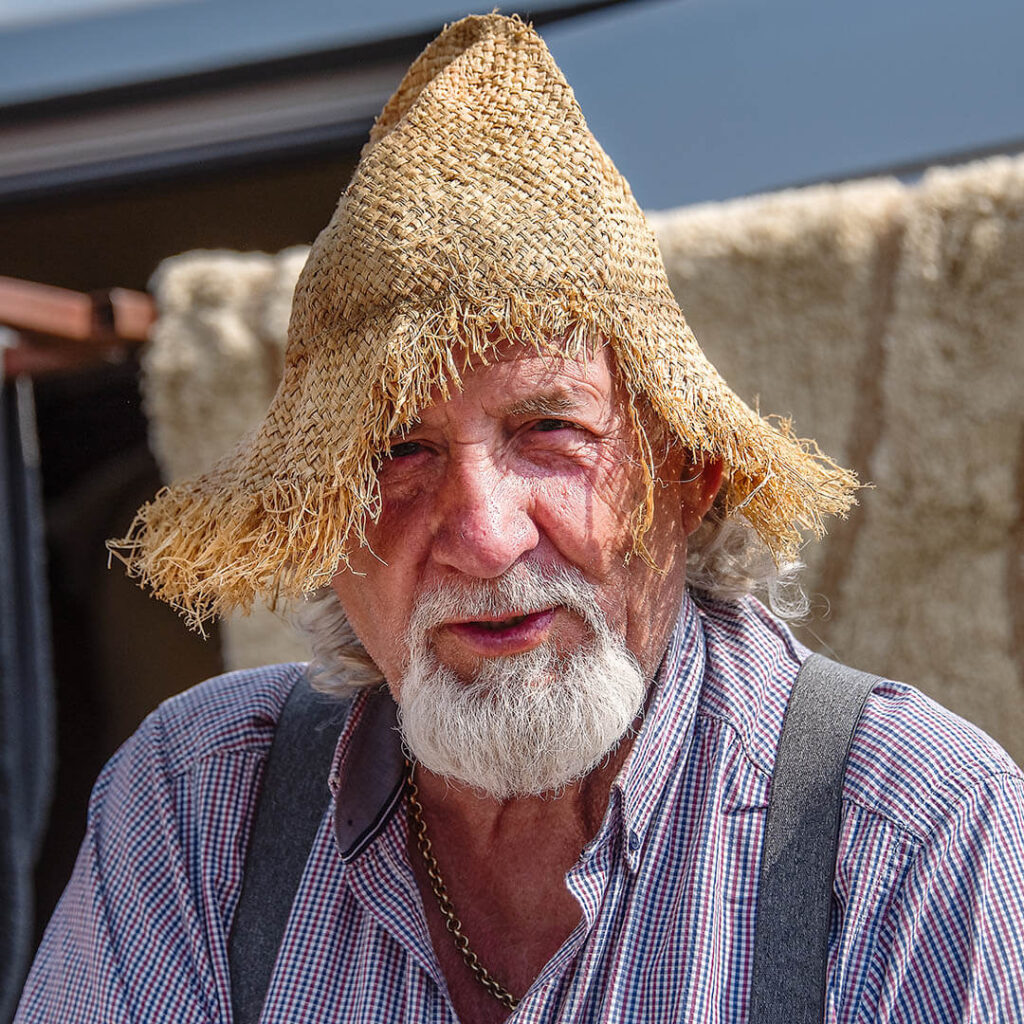 Portrait of an elderly man with a white beard, wearing a straw hat and a plaid shirt, at his trading stall at the Appleby Horse Fair.