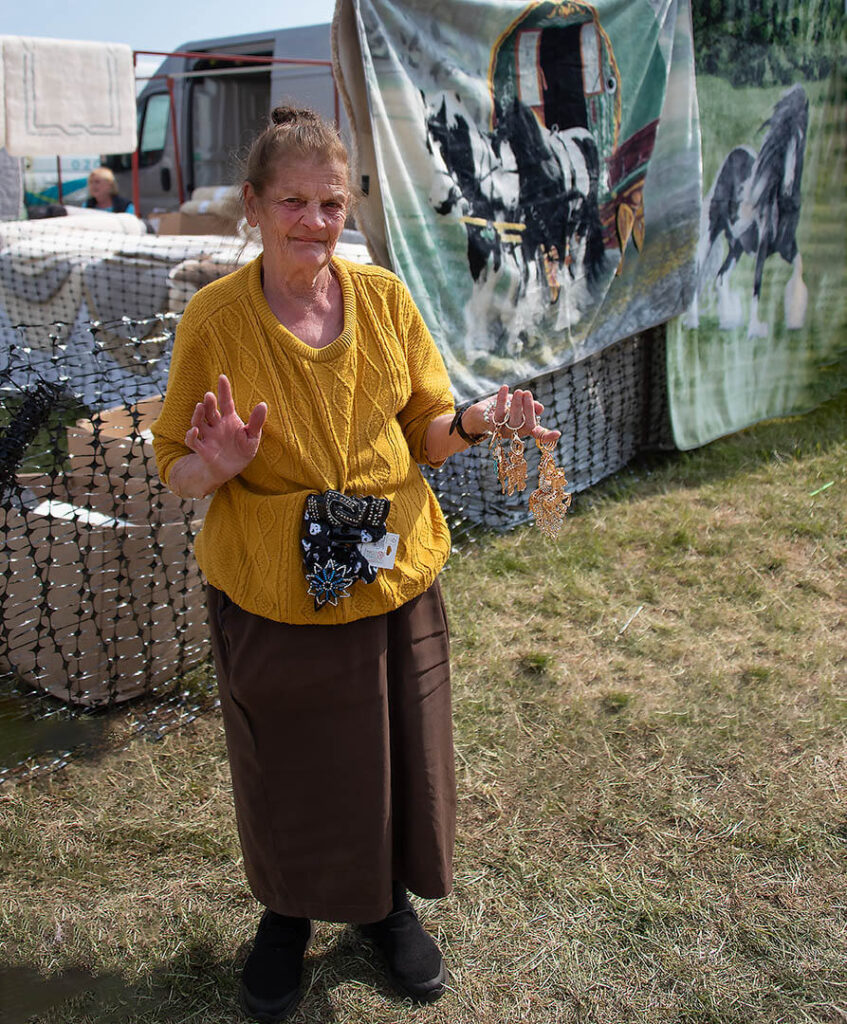 A woman named Rosie, wearing a yellow sweater and brown skirt, stands at her stall holding metal owl ornaments at the Appleby Horse Fair.