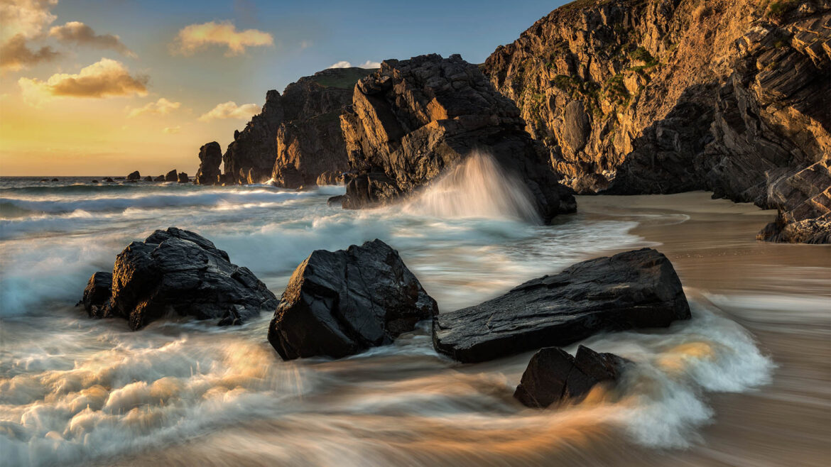 Dalmore Beach at sunset, golden light bathing the cliff faces, rocks, and sand while waves crash gracefully and create bubbling, streaked patterns.