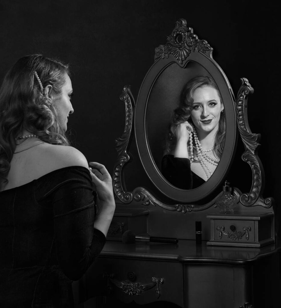 Elegant woman in a vintage black dress admiring her reflection in an ornate mirror, embodying classic Hollywood glamour.