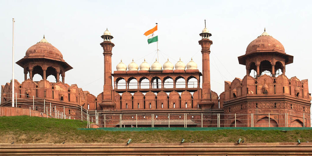 The iconic Red Fort, with its red sandstone walls, towers, and domes, under a clear sky, with the Indian flag flying proudly at its center