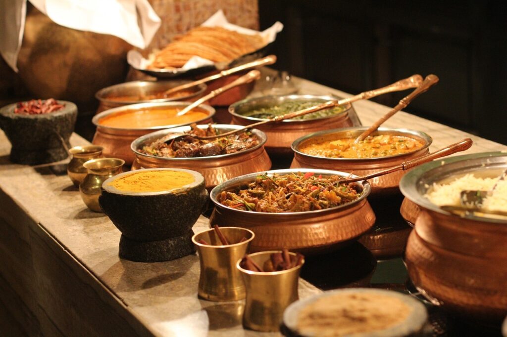 A traditional Indian buffet spread with a variety of dishes in copper bowls, including curries, rice, and bread, all laid out on a stone counter with serving spoons, under warm ambient lighting.