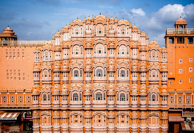 Hawa Mahal in Jaipur, showcasing its distinctive pink-red sandstone architecture with intricate latticework and patterned honeycomb windows, set against a blue sky.