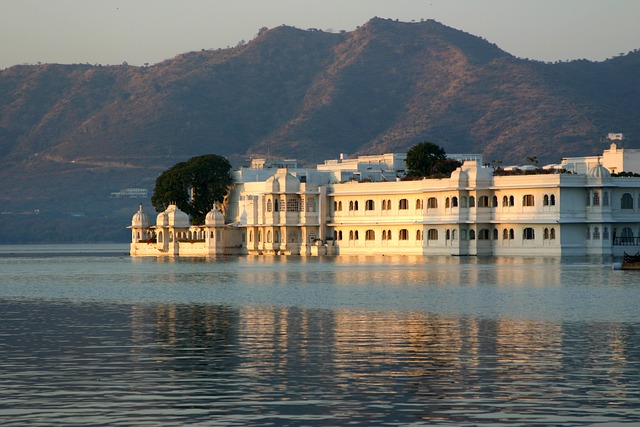 The Lake Palace of Udaipur, a white marble architectural wonder, floats on the still waters of Lake Pichola, with the Aravalli Mountains in the backdrop during a warm sunset.