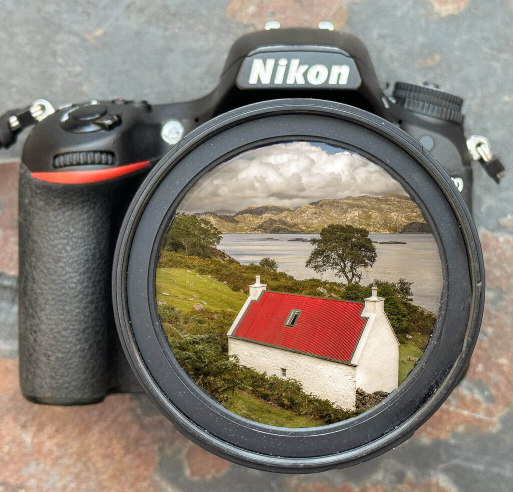 A Nikon camera positioned against a rustic backdrop, its lens capturing a serene landscape with a quaint white house with a red roof, nestled among verdant foliage beside a calm lake with distant mountains.