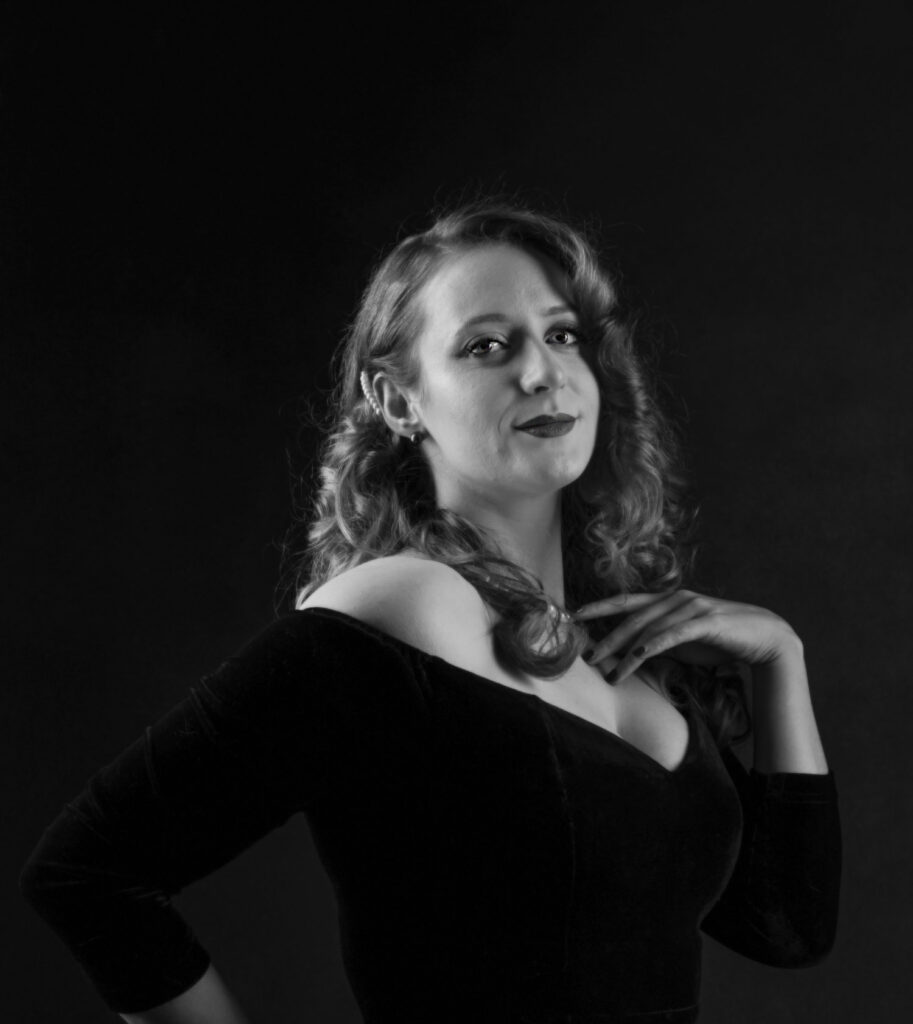 Black and white portrait of a woman with curly hair, wearing dark lipstick and a black velvet dress, posed with hands on her shoulders against a black background