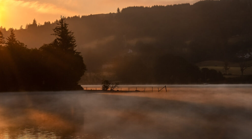 A tranquil Coniston lake at sunrise with a gentle mist hovering above the water, backlit by the golden hues of the early morning sun filtering through the trees and a quaint house on the hillside.