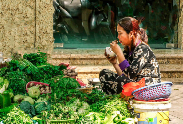 A woman enjoying a meal amid her market greens in Hanoi, Vietnam