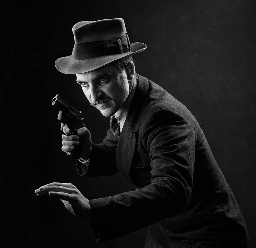 Monochrome photo of a man in a vintage outfit with a fedora hat, aiming a revolver towards the camera, his pose and expression intense and focused