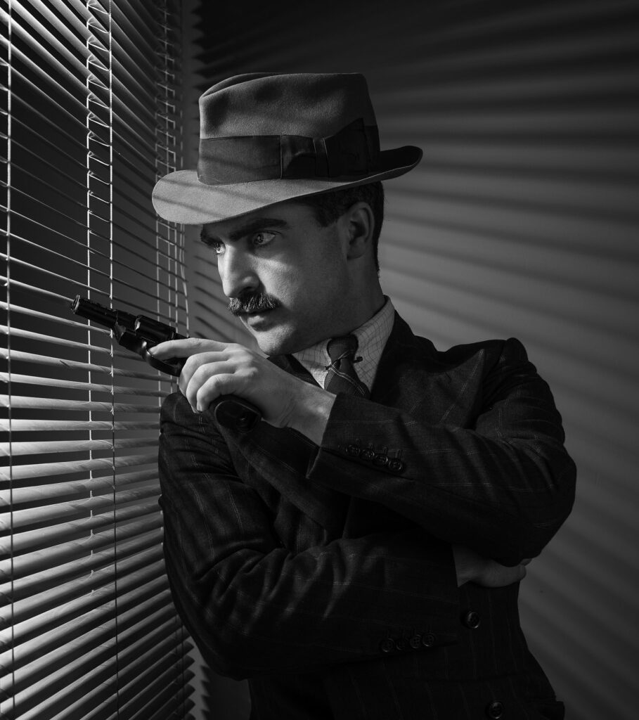 Black and white photo of a man dressed in 1940s attire, including a fedora hat, peering through window blinds with a revolver in his hand