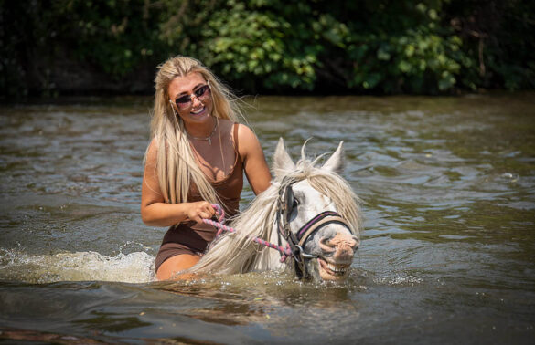 Blonde girl joyfully riding a white horse through the water at Appleby Horse Fair.