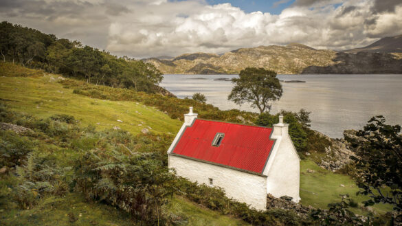 White cottage with a red roof set in lush green countryside by a lake.