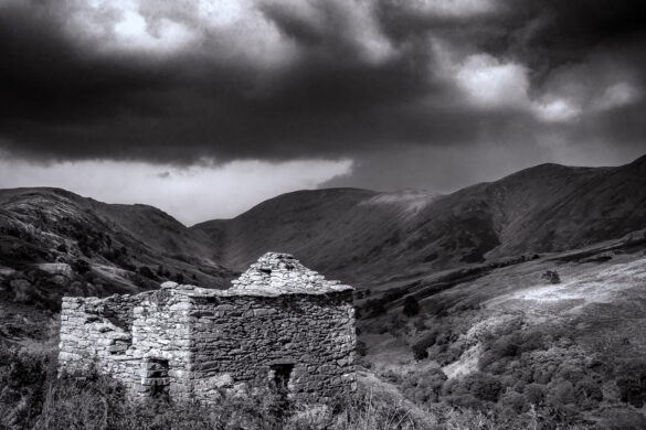 A crumbling stone hut set against a dramatic mountain backdrop in monochrome.